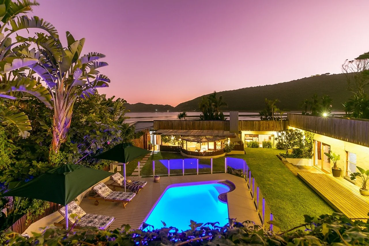 Lit garden, pool and lodge buildings above a lagoon at dusk on the Garden Route